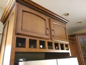 Upper kitchen cabinets with wooden doors, crafted by a skilled cabinet maker, feature six small square cubbyholes below, each holding a single bottle. The custom cabinets are above a stainless steel refrigerator under a ceiling with recessed lights.
