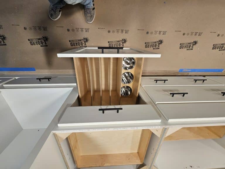 Overhead view of a custom cabinets installation featuring pull-out vertical dividers for baking sheets and built-in metal bins, surrounded by unfinished cabinetry and protective floor covering. A cabinet builder’s feet are visible at the top.